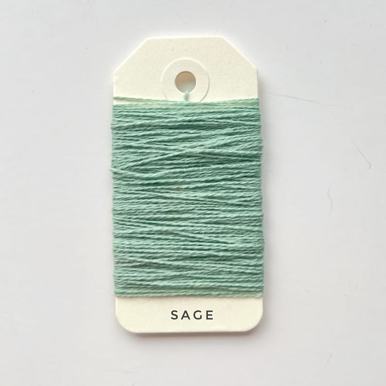 May include: A spool of sage green thread is wrapped around a cream-colored tag. The word "SAGE" is printed in black letters at the bottom of the tag. The thread is tightly wound, creating a textured appearance.