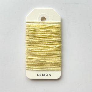 May include: A spool of lemon-yellow thread is wrapped around a cream-colored tag. The word "LEMON" is printed in black, centered at the bottom of the tag. The thread is tightly wound, creating a textured surface.