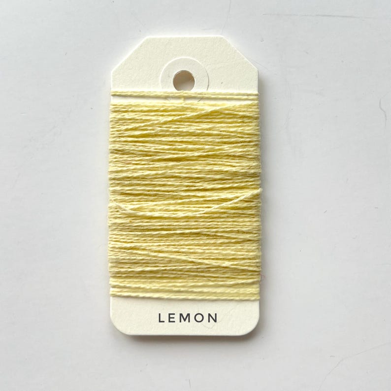 May include: A spool of lemon-yellow thread is wrapped around a cream-colored tag. The word "LEMON" is printed in black, centered at the bottom of the tag. The thread is tightly wound, creating a textured surface.