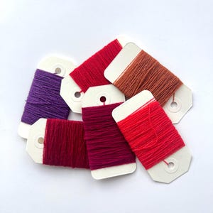 Puede incluir: Una colección de seis madejas de hilo de bordar enrolladas en tarjetas de cartón blancas. Los colores incluyen morado, rojo brillante, rojo oscuro, magenta, marrón anaranjado y un rojo coral vibrante, dispuestos en un montón superpuesto.