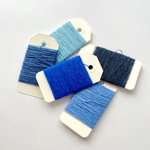 May include: Five spools of thread in shades of blue, from light sky blue to navy. Each spool is wrapped around a white tag with a small hole at the top. The threads are neatly wound, creating a visually appealing arrangement.