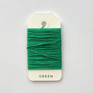 May include: A spool of bright green thread wrapped around a cream-colored tag. The tag has a hole at the top and the word "GREEN" printed at the bottom. The thread is tightly wound, creating a solid block of color.
