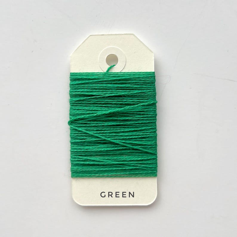 May include: A spool of bright green thread wrapped around a cream-colored tag. The tag has a hole at the top and the word "GREEN" printed at the bottom. The thread is tightly wound, creating a solid block of color.