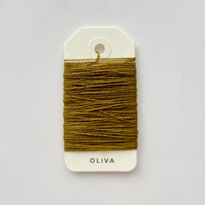 May include: A spool of olive-green thread is wrapped around a white tag. The tag has a hole at the top and the word "OLIVA" printed in black at the bottom. The thread is tightly wound, creating a textured appearance.