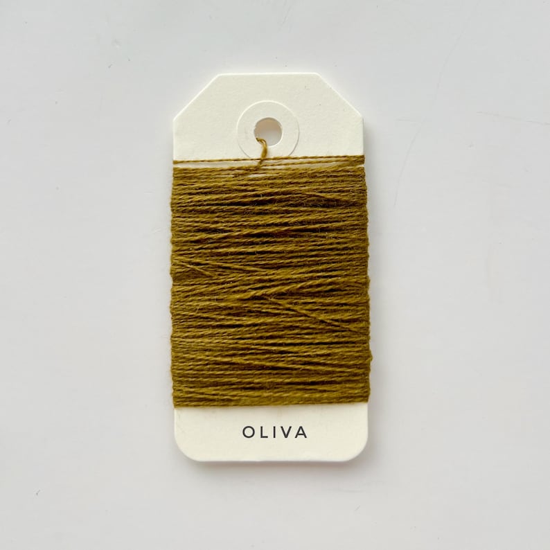 May include: A spool of olive-green thread is wrapped around a white tag. The tag has a hole at the top and the word "OLIVA" printed in black at the bottom. The thread is tightly wound, creating a textured appearance.