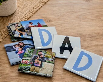 May include: A collection of square ceramic coasters with family photos and the word "DAD" spelled out in individual letter tiles. The "D" tiles have a light blue letter, and the "A" tile has a black letter. The coasters are arranged on a wooden surface.