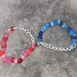 Könnte beinhalten: Zwei Perlenarmbänder mit den Wörtern "CHARACTER" und "CHARACTER" darauf. Das Armband links ist pink und rot mit Stern- und Herzperlen. Das Armband rechts ist blau mit Stern- und Smiley-Perlen.