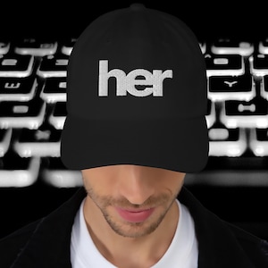 May include: Black baseball cap with the word "her" embroidered in white. The cap is worn by a person, with a blurred keyboard in the background.