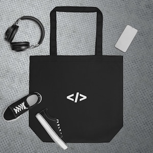 May include: Black tote bag with white angled brackets, "</>", printed on the front. The bag is on a silver background with a pair of black headphones, a black and white trainer, and a white smartphone.
