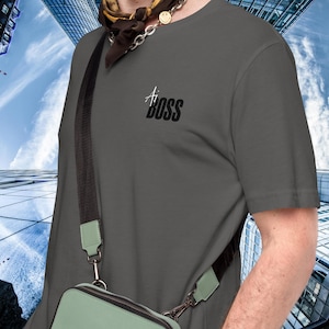 May include: A gray t-shirt with a black graphic that says "Ai Boss". The shirt is being worn by a person with a black and white patterned scarf around their neck. The person is also wearing a black crossbody bag.