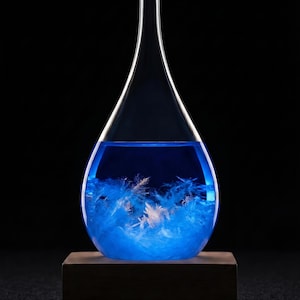 May include: A teardrop-shaped glass weather forecaster filled with blue liquid and feathery crystals, set on a wooden base. The glass has a long, slender neck and a rounded bottom. The crystals appear white and blue.