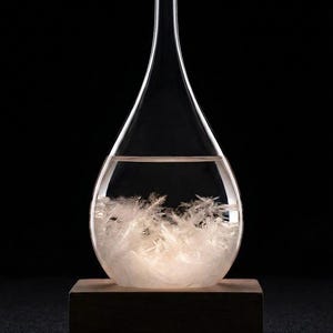 May include: A clear glass storm glass weather predictor shaped like a teardrop, filled with liquid and white crystalline formations, mounted on a rectangular dark wooden base against a solid black background.