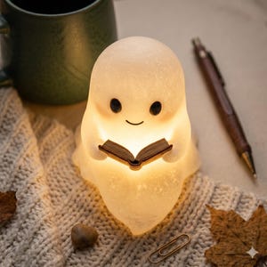 Op de afbeelding: Een oplichtend, wit spookvormig nachtlampje dat een klein open boek vasthoudt. De lamp heeft zwarte ogen en een lachende mond. Het staat op een crèmekleurig gebreid oppervlak, met een pen en een mok op de achtergrond.