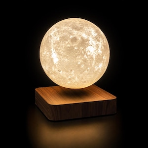 May include: A levitating moon lamp featuring a spherical, textured lunar surface that emits a warm, soft glow. The sphere is suspended above a square, polished wooden base with a natural grain finish, creating a floating effect in the dark.