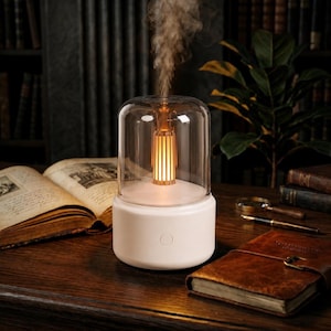 Peut inclure: Un diffuseur d'huiles essentielles cylindrique blanc avec un dôme en verre transparent. À l'intérieur du verre, un élément lumineux vertical central émet une lueur orange chaude tout en libérant un jet de brume visible par le haut.