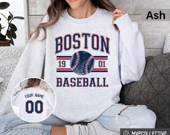 May include: A gray sweatshirt with the text "BOSTON BASEBALL" in navy blue and red. A baseball with red stitching is centered above the text. The numbers "19" and "01" are on either side of the baseball. The sweatshirt is shown on a model wearing blue jeans.