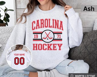 Puede incluir: Una sudadera gris con el texto "CAROLINA HOCKEY" en rojo y negro. La sudadera tiene un gráfico de palo de hockey y disco con los números 19 y 71. La sudadera se muestra en una modelo que lleva pantalones vaqueros azules.