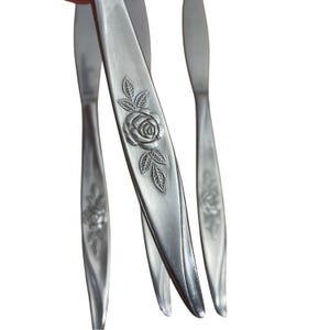 May include: Close-up of several stainless steel butter knives. Each knife features a floral rose design on the handle. The knives are silver and have a smooth, reflective surface. The handles are elongated and slightly curved.