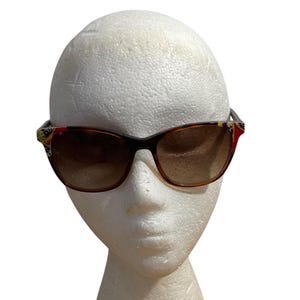 May include: Brown sunglasses with a dark frame and gradient lenses. The temples feature colorful decorative accents with red, yellow, and green patterns. The sunglasses are displayed on a white mannequin head.
