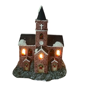 May include: A miniature brick church figurine featuring a tall central steeple with a white cross, dark shingled roofs topped with white snow, and arched windows glowing with warm interior light. The structure includes three front doors.