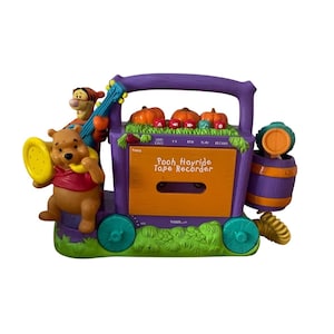May include: A purple and orange Winnie the Pooh Hayride tape recorder featuring a Tigger figure holding a blue guitar and a Pooh figure holding a yellow horn. The device includes a cassette compartment, control buttons, and pumpkin accents.