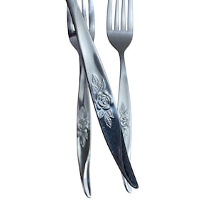 May include: Close-up of three silver forks with a floral rose design on the handles. The forks are arranged in a slightly overlapping pattern against a white background. The tines of the forks are visible.