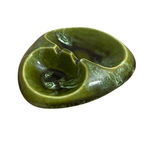 May include: An irregular, rounded ceramic dish featuring a glossy olive green glaze with darker brown, mottled accents. The surface has a smooth, reflective finish with two distinct, shallow recessed sections separated by a central ridge.