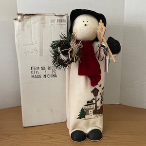 May include: A snowman figurine with a black top hat, red scarf, and a sign that reads "Let it Snow". The snowman holds a green wreath and has a painted face. The base of the snowman has a winter scene with a snowman and birdhouse.