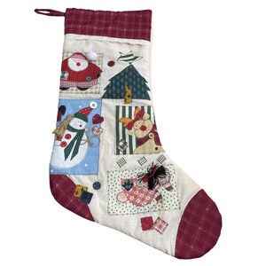 May include: A Christmas stocking with a patchwork design featuring festive images. The stocking has a burgundy plaid cuff and toe, with appliqued squares of Santa, a snowman, and a reindeer. The background is cream-colored with decorative stitching.