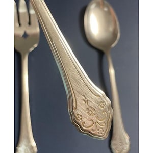 May include: Close-up of antique silverware, including a fork, spoon, and knife. The silverware has a silver finish with ornate floral detailing on the handles. The fork has a star-shaped cutout.