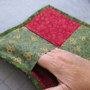 May include: A square quilted fabric potholder featuring a patchwork design with alternating sections of olive green fabric with a small yellow and red floral pattern and a bright red fabric with a white and orange circular floral print.