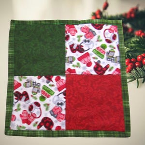 May include: A square fabric coaster featuring a four-patch design with alternating blocks of solid dark green fabric, solid red fabric, and white fabric printed with a pattern of red and green winter mittens, hats, and earmuffs, all bordered by green plaid.
