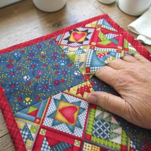 May include: A square quilted pot holder featuring a patchwork design with blue fabric printed with small red flowers and yellow paw prints, alongside blocks displaying red hearts, green geometric patterns, and a red fabric border trim.
