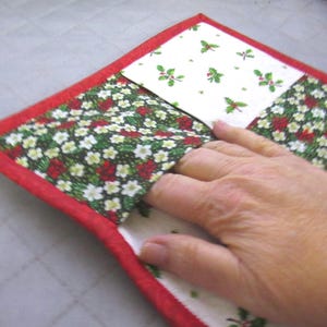 May include: A quilted fabric potholder featuring a festive holiday pattern with white, green, and red holly berries and leaves. The square item is finished with a bright red fabric binding along the edges and includes a pocket for hand insertion.