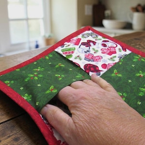 May include: A quilted fabric pot holder featuring a red border and two distinct patterns. One section displays a green fabric with a holly leaf and berry print, while the other section shows a white fabric with a colorful winter mitten and hat print.
