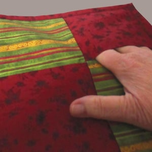 May include: A quilted fabric square featuring alternating panels of deep red fabric with a dark floral pattern and green fabric with horizontal stripes in yellow, orange, and red. The square is stitched with a border and has a soft, padded texture.