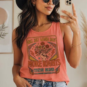 Könnte beinhalten: Korallenfarbenes Tanktop mit dem Text "GIRLS JUST WANNA HAVE MORE RIGHTS THAN GUNS" in einem Retro-Design. Die Grafik zeigt eine Hand, die Blumen, Blitze und Pistolen hält. Das Tanktop ist ärmellos und aus weichem Material.