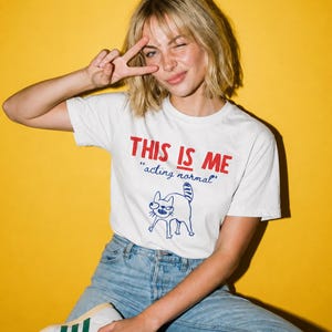 May include: A white short-sleeve t-shirt featuring the red text THIS IS ME, the blue cursive text acting normal, and a blue line drawing of a cat with a striped tail. The shirt is paired with light blue denim jeans and white sneakers.