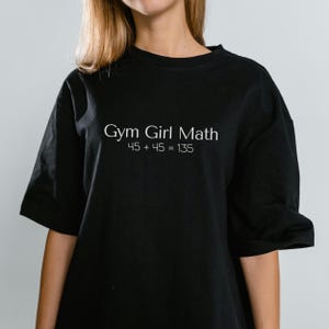May include: Black t-shirt with the white text "Gym Girl Math" and the incorrect equation "45 + 45 = 135". The shirt has short sleeves and a crew neck.