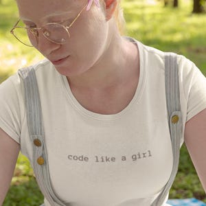 Könnte beinhalten: Cremefarbenes T-Shirt mit dem Aufdruck "code like a girl". Das Shirt wird mit grauen Hosenträgern und einem passenden Rock getragen. Eine Blume ist hinter dem Ohr platziert.