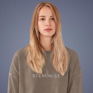 Könnte beinhalten: Ein taupefarbenes Sweatshirt mit dem weißen Aufdruck "STEMINIST" und einer kleinen botanischen Illustration. Das Sweatshirt wird von einer Person mit langen blonden Haaren vor blauem Hintergrund getragen.