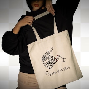Könnte beinhalten: Cremefarbene Tragetasche mit einer schwarzen Grafik eines Laptops und einer Hand mit rotem Nagellack. Der Text "Freak in the Sheets" befindet sich unter der Laptop-Illustration. Die Tasche hat zwei Griffe.