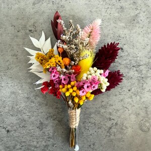 Op de afbeelding: Een kleurrijk boeket van gedroogde bloemen met witte, gele, roze, rode en bordeauxrode bloemen. De stelen zijn samengebonden met touw en een kleine zilveren speld houdt de bloemen bij elkaar. Een gevarieerde en getextureerde bloemenpresentatie.