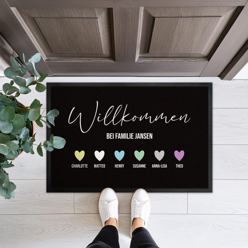 May include: Black rectangular welcome mat with the word "Willkommen" in white script. Below, it reads "Bei Familie Jansen" and features six colorful hearts with names. A person is standing on the mat.