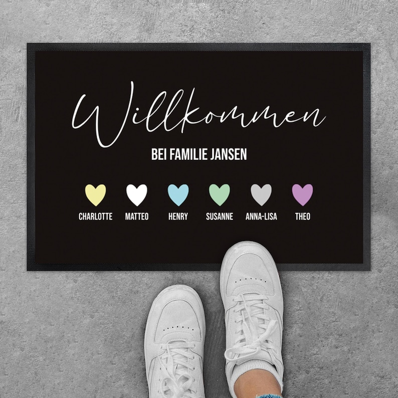 May include: Black welcome mat with the word "Willkommen" in white script, followed by "Bei Familie Jansen". Below are six colored hearts with names. White sneakers are visible at the bottom.