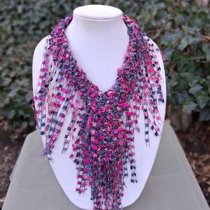 May include: A handmade necklace displayed on a white mannequin. The necklace features a vibrant mix of pink, blue, and purple fabric strips, creating a textured, fringed design. The necklace is a statement piece, perfect for adding a pop of color.