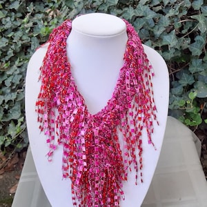May include: A pink and red beaded necklace with a fringe design. The necklace is made of small, square beads and has a long, flowing fringe.