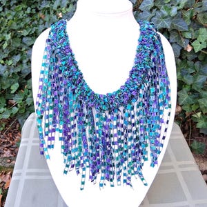 K&ouml;nnte beinhalten: Handgefertigte Halskette auf einer wei&szlig;en B&uuml;ste. Die Kette besteht aus gewebten B&auml;ndern in Blau-, T&uuml;rkis- und Lilat&ouml;nen. Die Fransen verleihen dem Design Textur und Farbe. Ein einzigartiges Schmuckst&uuml;ck.