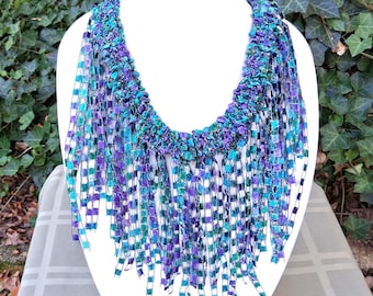 May include: A handmade necklace displayed on a white mannequin. The necklace features a woven design with a mix of blue, teal, and purple ribbons. The fringe hangs down, creating a textured and colorful statement piece.