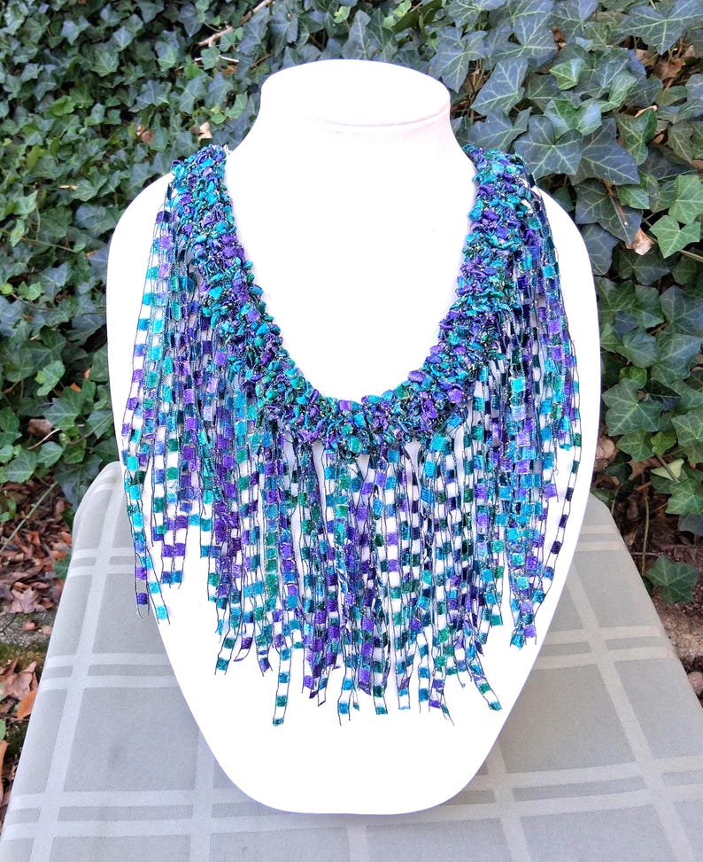 K&ouml;nnte beinhalten: Handgefertigte Halskette auf einer wei&szlig;en B&uuml;ste. Die Kette besteht aus gewebten B&auml;ndern in Blau-, T&uuml;rkis- und Lilat&ouml;nen. Die Fransen verleihen dem Design Textur und Farbe. Ein einzigartiges Schmuckst&uuml;ck.